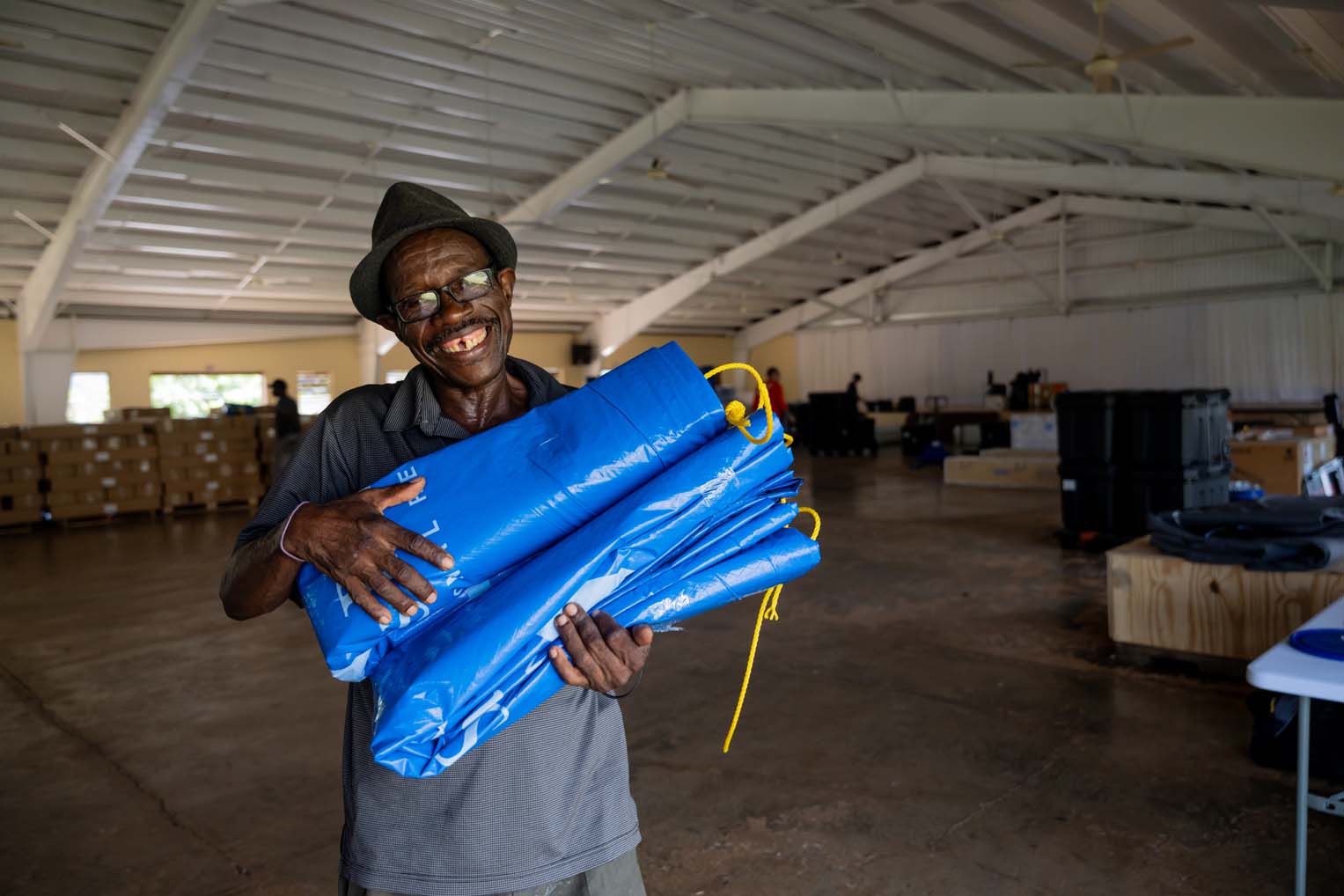Smiling Jamaican church leader holding blue tarp supplies inside a Samaritan’s Purse distribution warehouse after Hurricane Melissa.