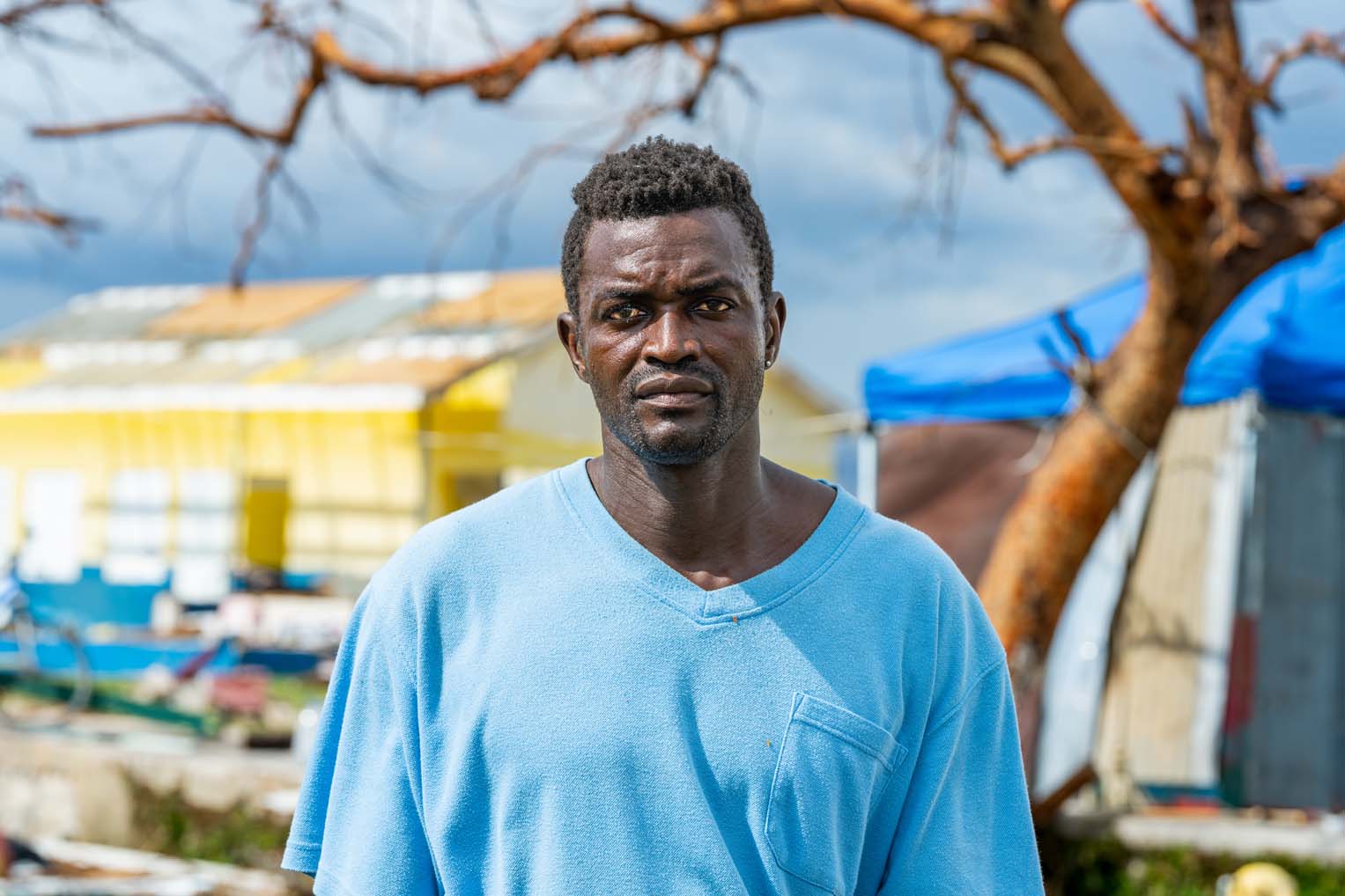 Close-up portrait of a man standing in front of storm-damaged buildings and trees in Black River, Jamaica, after Hurricane Melissa.