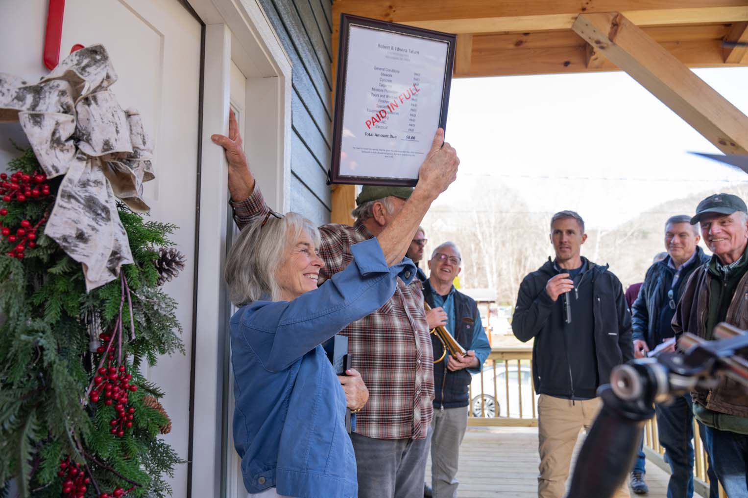 Edwina Tatum lifts the framed “Paid in Full” invoice on the porch of their newly rebuilt home as volunteers and guests look on.