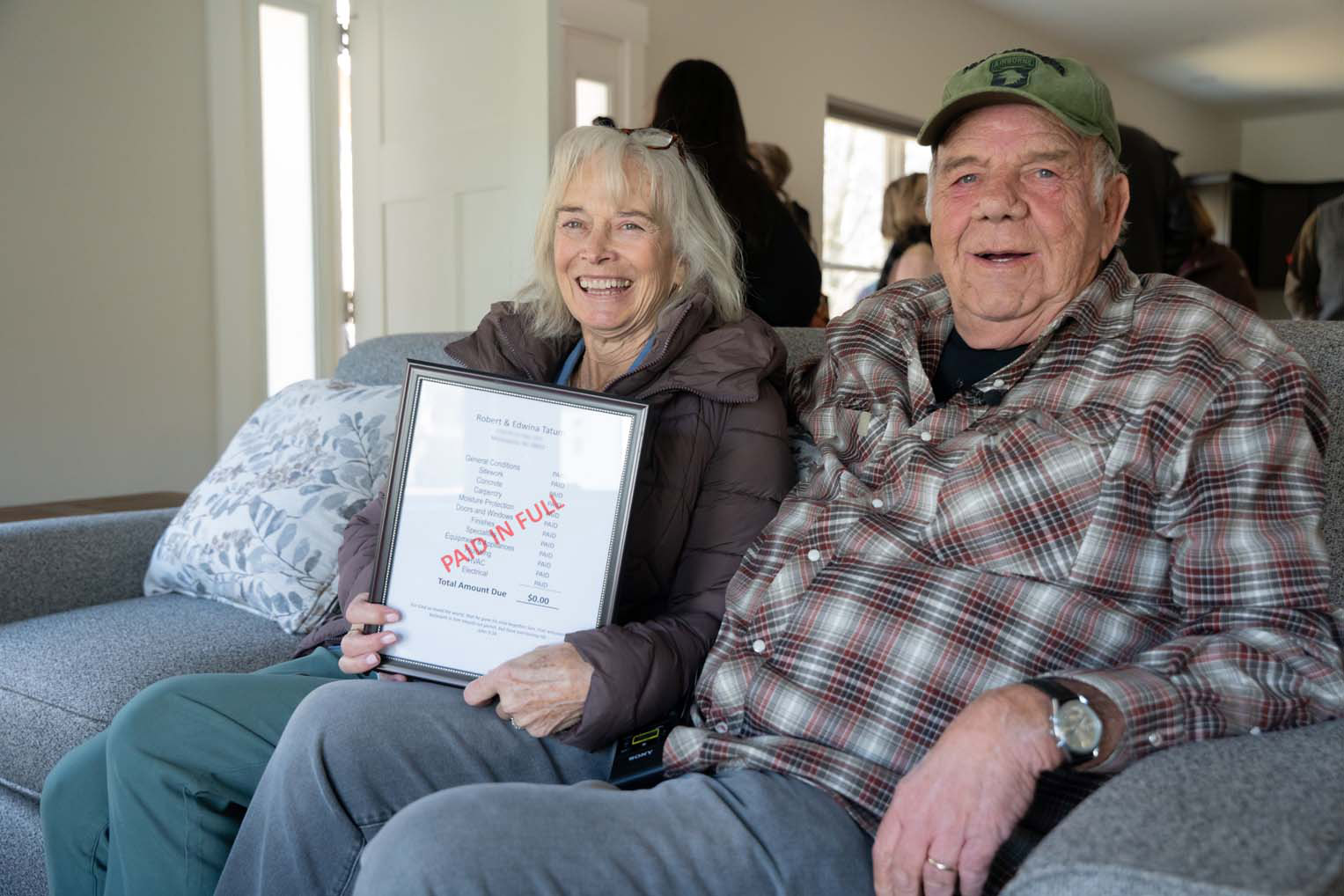 Bob and Edwina Tatum sit inside their new living room holding the framed “Paid in Full” document presented during their home dedication.