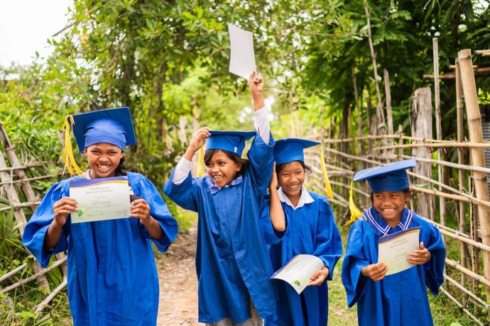 Four Cambodian girls in graduation gowns hold certificates of completion from The Greatest Journey