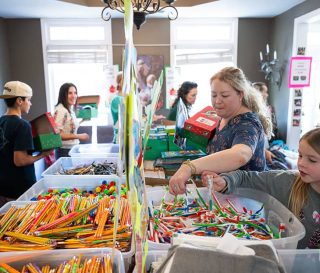 Adults and children packing shoeboxes with gifts from bins on a table in a house.