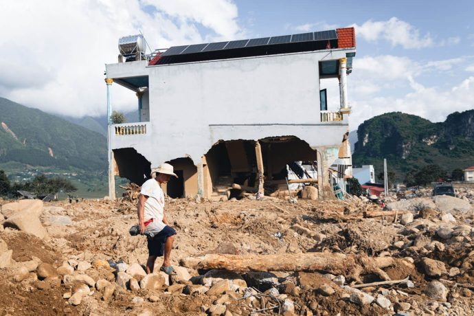 Man walks through field of boulders with partially destroyed home in background